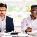 Two men in an office environment working at laptops on the same desk